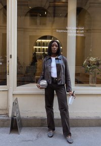 Braune Lederjacke, weißes Oberteil, dunkle Hose, schwarz-weiße gemusterte Schuhe. Hält eine glitzernde Tasche und ein Getränk mit einem Strohhalm.