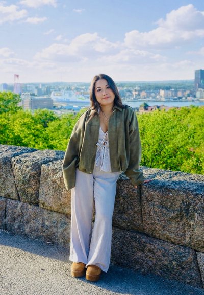 Mujer joven con chaqueta verde y pantalones blancos apoyada en un muro de piedra, con la ciudad y el río al fondo bajo un cielo azul.