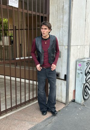 Joven con chaleco de cuero negro, camisa de manga larga a rayas rojas y negras, vaqueros negros y zapatos negros, de pie junto a una puerta de hierro y una pared de cemento.