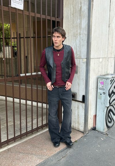 Joven con chaleco de cuero negro, camisa de manga larga a rayas rojas y negras, vaqueros negros y zapatos negros, de pie junto a una puerta de hierro y una pared de cemento.