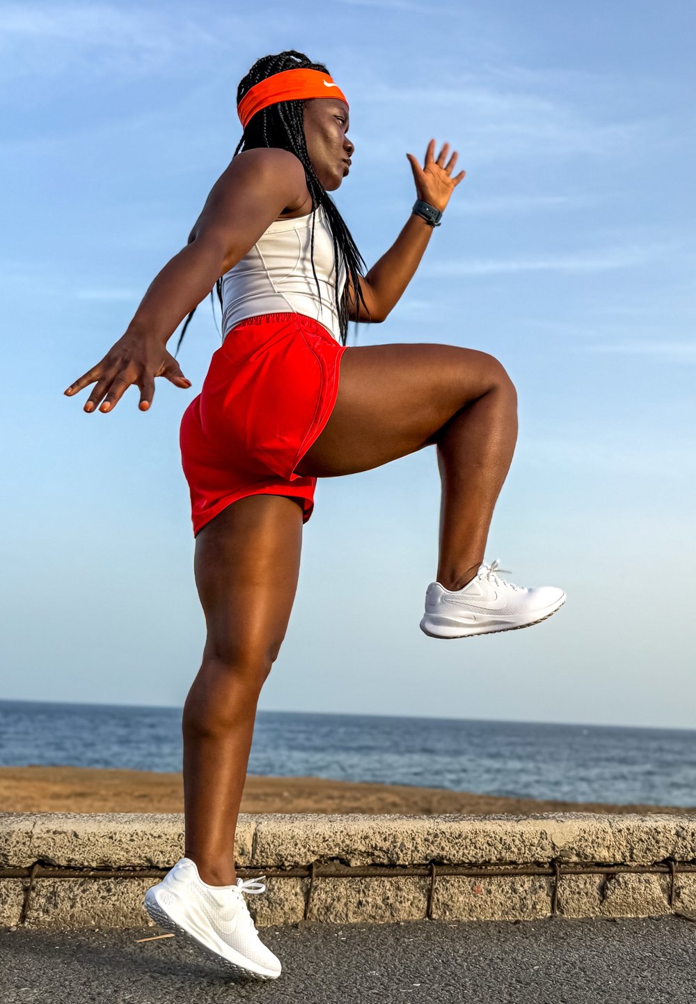 Sportowa osoba w białej koszulce bez rękawów i czerwonych szortach, nosząca pomarańczową opaskę na głowie i białe sneakersy, w trakcie biegu wzdłuż nadmorskiej ścieżki.