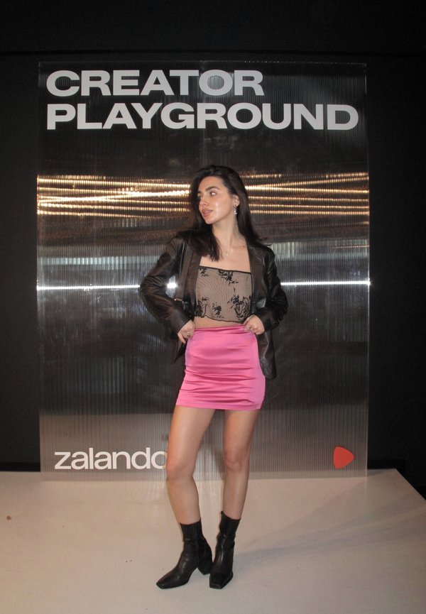 Tânără cu jachetă neagră, top din dantelă, fustă roz și cizme negre, pozând în fața unui panou cu inscripția „CREATOR PLAYGROUND” și „zalando.”