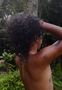 Personne aux cheveux bouclés et foncés, avec des tatouages sur le bras gauche et le côté, debout torse nu dans une forêt verdoyante, les mains levées derrière la tête.