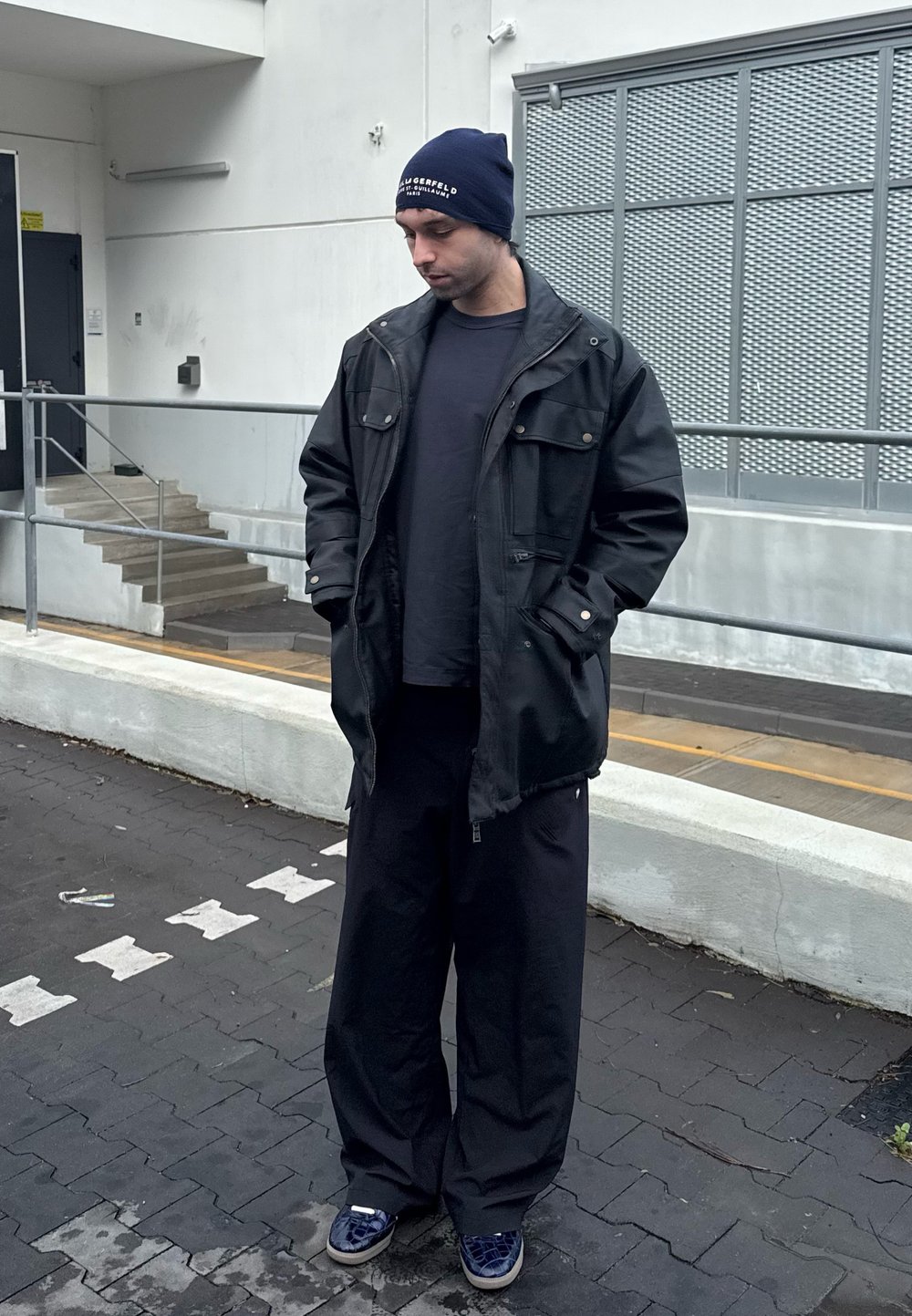 Crna kožna jakna, tamnoplava beanie s logom, tamna majica i prevelike crne hlače. Obuća uključuje plave patike s uzorkom.