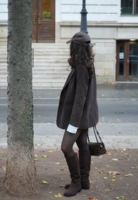 Smeđi široki sako, bijele rukave košulje, smeđe duge čizme, tamna kapa i mala crna torbica sa zlatnim zakovicama. Stoji pored drveta.