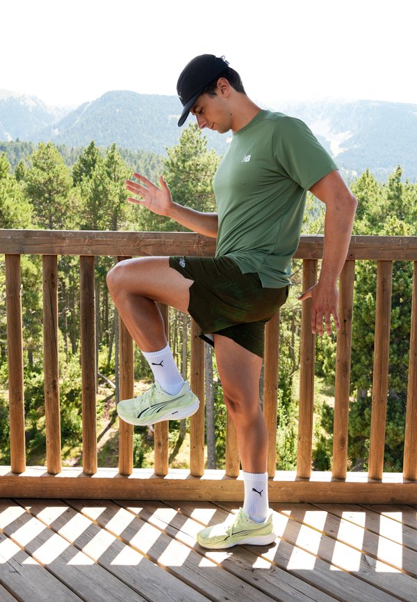 Homme en tenue de sport et casquette étirant la jambe sur un balcon en bois avec vue sur la forêt et la montagne en arrière-plan.