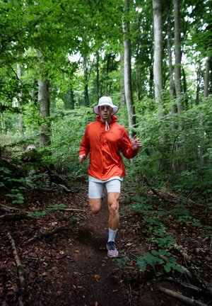 Corridore con una giacca leggera arancione, pantaloni corti bianchi e scarpe da ginnastica grigie, che percorre un sentiero nel bosco circondato da vegetazione verde e alberi.