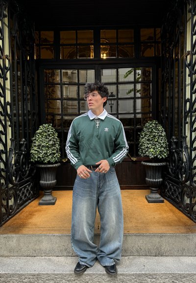 Polo de manga larga verde con rayas blancas, jeans anchos azules, cinturón negro, anillos plateados y gafas, frente a una puerta decorativa.