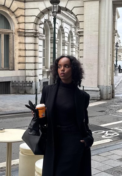 Mujer con cabello rizado que lleva un abrigo negro y guantes, sostiene una taza de café mientras está de pie en una calle de la ciudad con edificios históricos de piedra blanca.