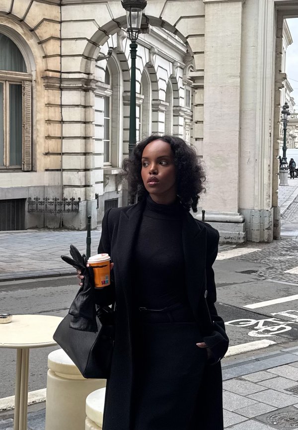 Femme aux cheveux bouclés portant un manteau noir et des gants, tenant une tasse de café, debout dans une rue de la ville avec des bâtiments historiques en pierre blanche.