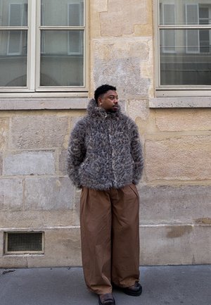 Chaqueta marrón de piel sintética con cuello alto, cremallera frontal y bolsillos grandes; combinada con pantalones marrones de corte holgado y zapatos negros.