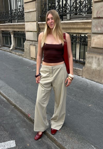 Una mujer está de pie en una calle, vistiendo una blusa ajustada marrón y pantalones anchos beige, sosteniendo una bolsa roja, con zapatos rojos de punta y accesorios en la muñeca.