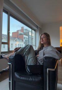 Chaise en cuir noir avec accoudoirs en chrome, une personne en pull gris et pantalon en denim, assise près de grandes fenêtres offrant une vue urbaine.
