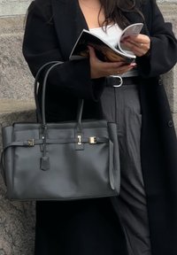 Crna kožna torba s zlatnim detaljima, pravokutnog oblika, s ravnim ručkama i glatkom teksturom. Drži časopis, nosi se uz sive hlače.