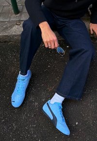 Personne portant des baskets Puma bleues, un jean foncé et un pull noir assise par terre en tenant des lunettes de soleil à la main.