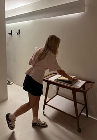 Holzservierwagen mit weißen Regalen; ein Regal ist mit Büchern gestapelt. Person in einem pink gestreiften Shirt und dunklem Rock, die nach etwas greift.