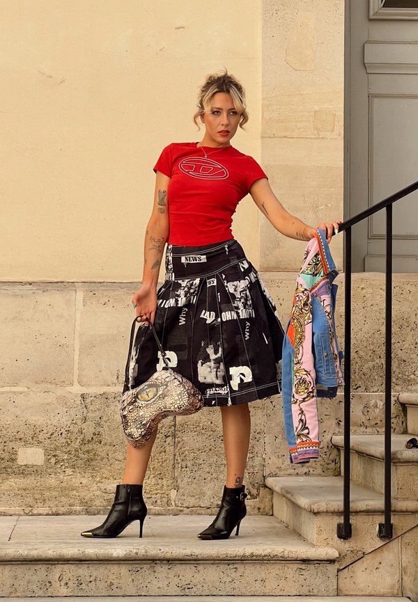 Femme portant une chemise rouge et une jupe noire imprimée de journaux, tenant une veste à motifs et un sac à main, debout sur des marches en pierre près d'une rambarde.