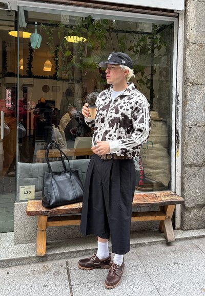 Chaqueta de algodón con diseño de vaca, pantalones cortos anchos negros, zapatos marrones con calcetines blancos y un bolso negro. De pie fuera de un café.