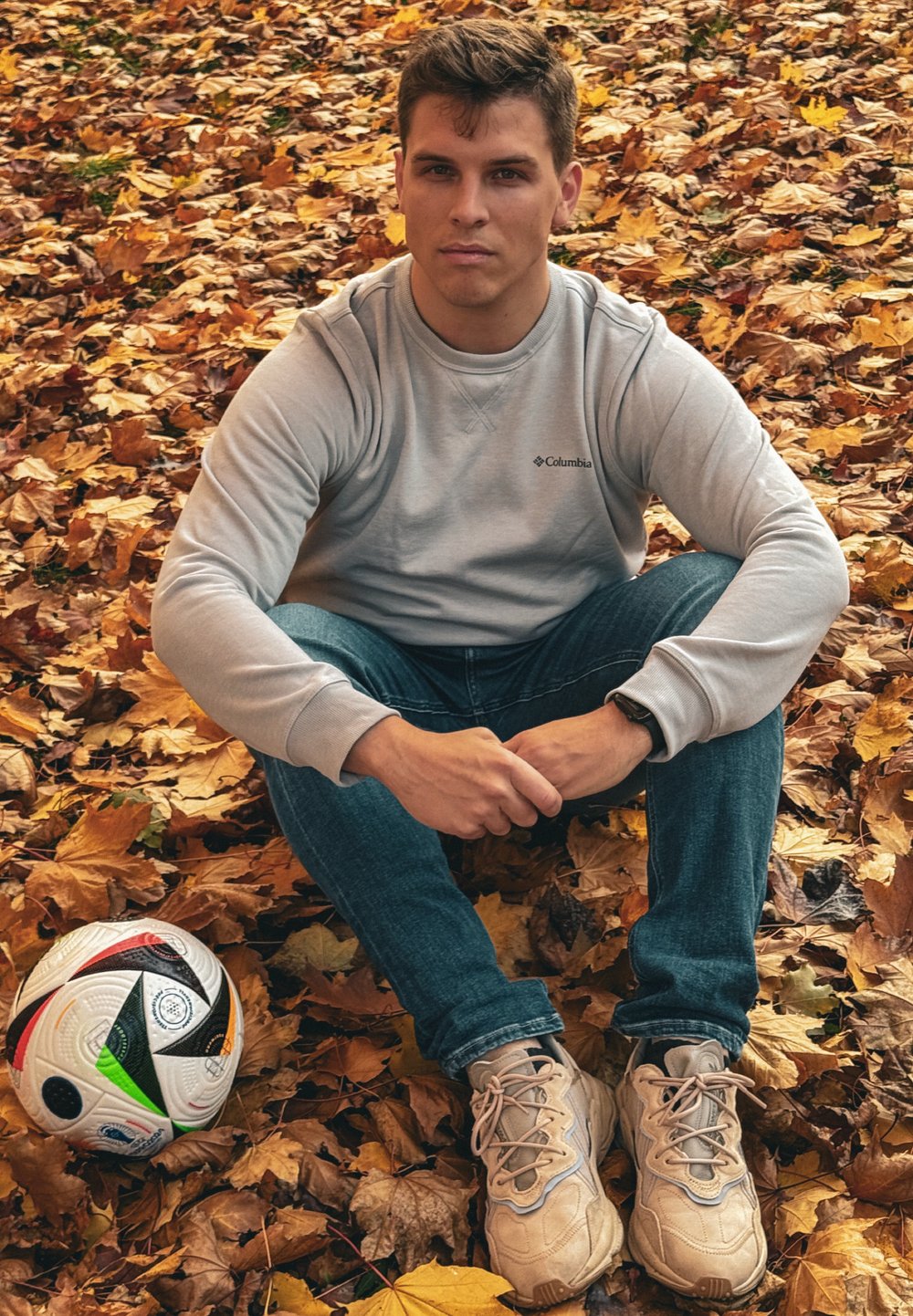 Γκρι φούτερ με στρογγυλή λαιμόκοψη, μπλε τζιν και μπεζ αθλητικά παπούτσια. Μια μαύρη και άσπρη μπάλα ποδοσφαίρου είναι τοποθετημένη στο έδαφος ανάμεσα σε πορτοκαλί και καφέ φύλλα.