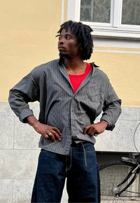 Szara, oversize koszula w paski z czerwoną koszulką pod spodem, zestawiona z ciemnoniebieskimi dżinsami. Na palcach widoczne charakterystyczne pierścionki, swobodnie oparta o ścianę.