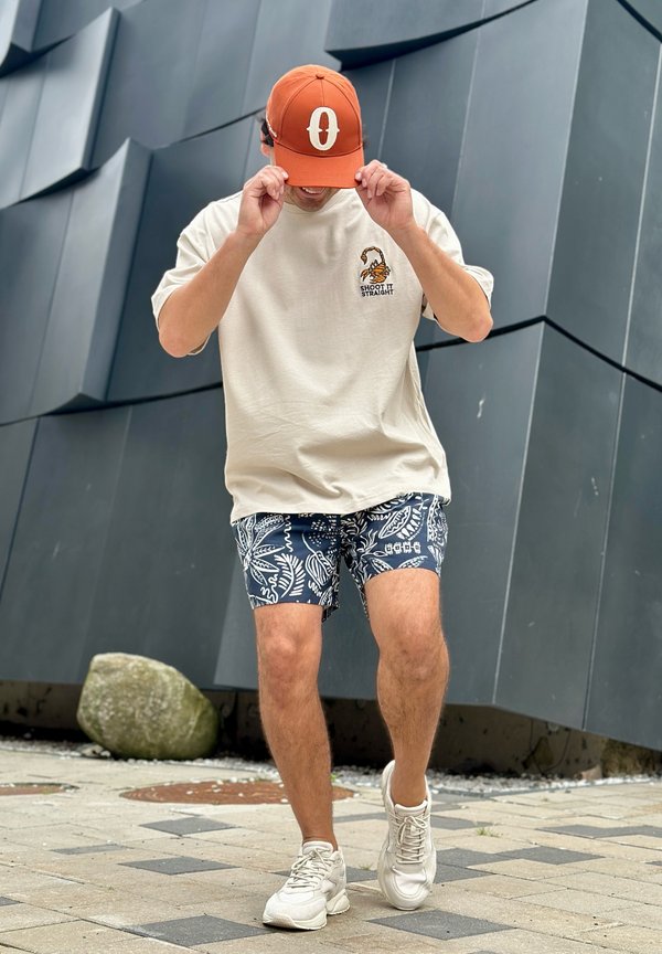 Homme portant une casquette orange, un t-shirt beige avec un motif de scorpion, un short bleu marine à motifs et des baskets blanches, debout devant un mur gris texturé.