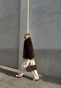 Rjava puhasta bunda, smetana široke hlače, svetlo rjave čevlje, drži temno rjav ročni torbico z zlatimi detajli, hodi ob teksturirani sivi steni.