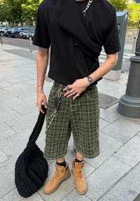 Fekete, rövid ujjú póló egy kötött mellénnyel, zöld kockás cargo sort, bézs munkanadrágot és egy fekete textúrájú táskát visel. Számos gyűrű és lánc.