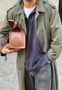 Καφετί δερμάτινη τσάντ α με ορθογώνιο σχήμα και λεία υφή, κρατημένη από ένα άτομο που φοράει παλτό ελαιώδους πράσινου χρώματος και ναυτικό πόλο μπλουζάκι.