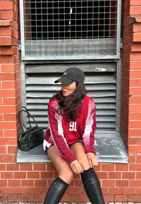 Bordo sweatshirt z belimi detajli in številko 91, v kombinaciji z črnimi čevlji do kolen. Črna torbica leži ob modelu na zidaku.