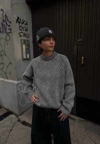 Knitted grey and black striped jumper with a high neckline, paired with a grey beanie featuring a logo. Loose dark trousers and multiple rings.