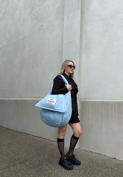 Bolso acolchado de color azul claro con forma redondeada, textura acolchada y una etiqueta blanca rectangular. Modelo vistiendo un atuendo negro y gafas de sol.