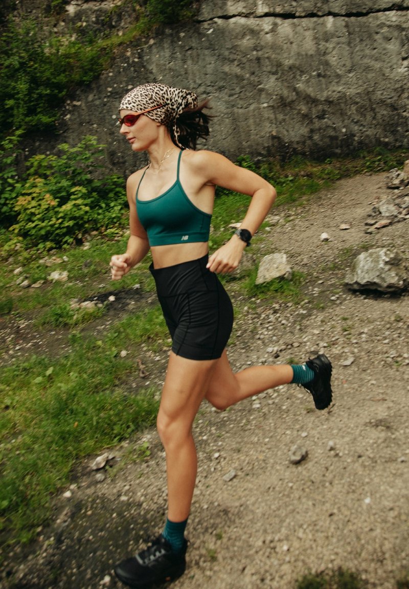 Femme portant un foulard léopard sur la tête et un soutien-gorge de sport vert courant sur un sentier rocailleux à côté d'un mur en pierre et de la verdure.