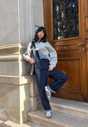 Joven mujer con suéter gris y peto vaquero apoyada en un muro de piedra junto a una gran puerta de madera con hierro decorativo, con un pie levantado sobre un escalón.