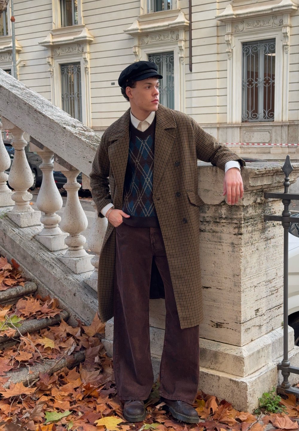 Long brown check coat over a dark jumper with a diamond pattern, wide brown trousers, black cap, and black shoes; autumn leaves on the ground.