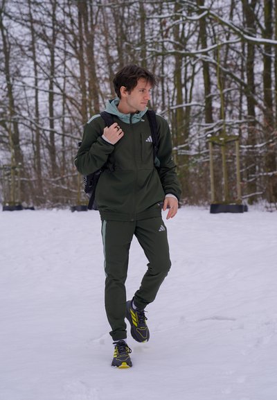 Hombre con chándal verde y zapatos con detalles amarillos caminando por un bosque cubierto de nieve, llevando una mochila negra y mirando hacia la derecha.