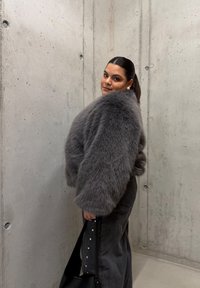 Veste courte en fausse fourrure grise à manches larges, portée sur un pantalon en denim foncé orné d'accents décoratifs en perles, sur fond de mur en béton.