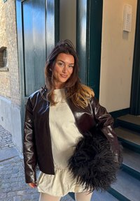 Hnědá kožená bunda s lesklým povrchem, nošená přes světlý top; drží černý měkký doplněk. V pozadí kamenná zeď a schody.