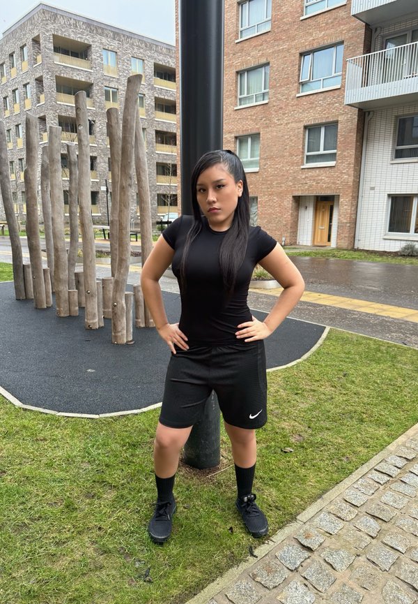 Camiseta y pantalones cortos deportivos negros con un logo blanco de Nike, de pie sobre una superficie texturizada con hierba y estructuras de madera al fondo.