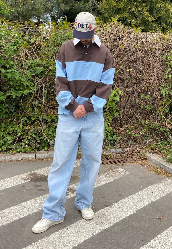 Mann, der ein braun-hellblau gestreiftes Langarmhemd, weite hellblaue Jeans, weiße Sneakers und eine Baseballkappe trägt und auf einem Zebrastreifen steht.