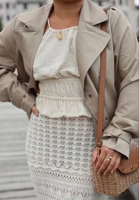 Beige trench coat over a white textured halter top, teamed with a white lace skirt that has a diamond pattern. Brown crossbody bag adorned with studs.