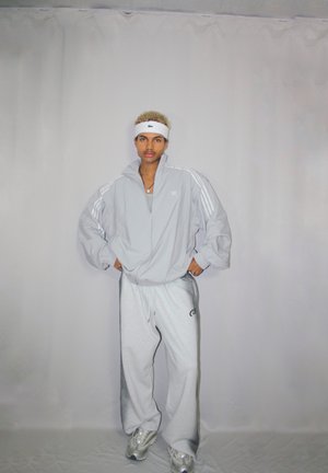 Chaqueta de chándal gris con franjas blancas, combinada con pantalones de chándal grises que llevan un logo. El modelo lleva una cinta blanca en la cabeza y zapatillas.