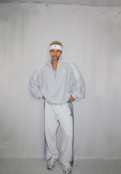 Chaqueta de chándal gris con franjas blancas, combinada con pantalones de chándal grises que llevan un logo. El modelo lleva una cinta blanca en la cabeza y zapatillas.