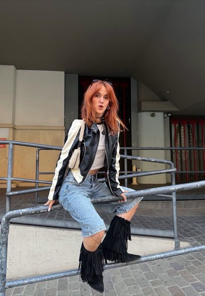 Chaqueta de cuero en blanco y negro, top blanco corto, pantalones cortos de mezclilla y botas negras con flecos. Sentada en una barandilla de metal contra una pared texturizada.