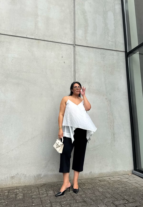 Mujer con top blanco sin mangas de varias capas, pantalones negros cortos, tacones negros, sosteniendo un pequeño bolso blanco, ajustándose las gafas, de pie junto a una pared de hormigón.