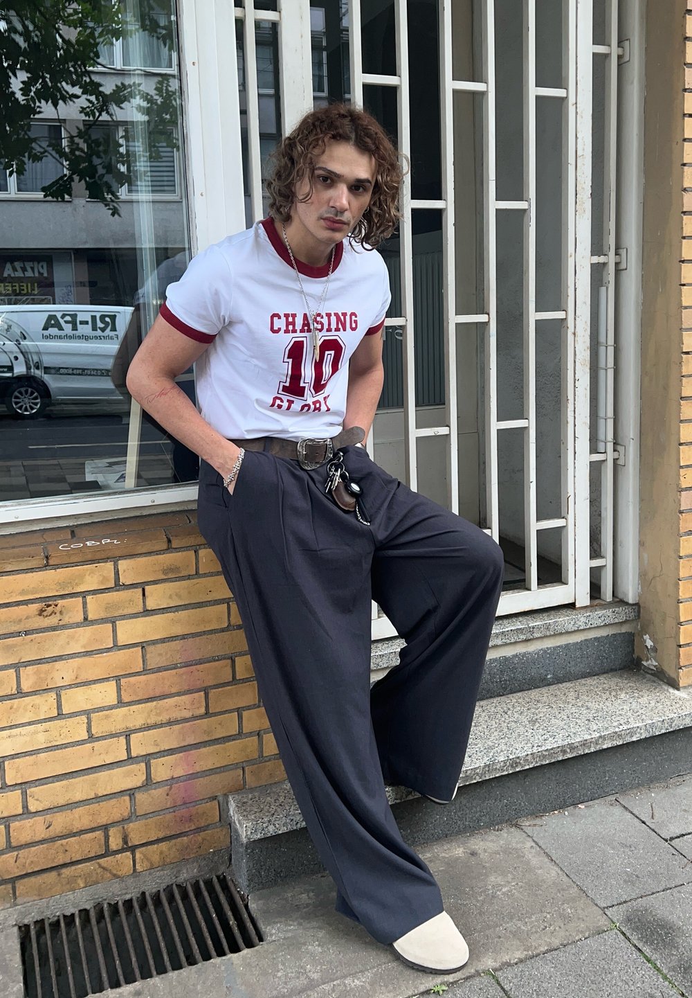 White t-shirt with red accents and the text "CHASING 10", teamed with dark wide-leg trousers and light-coloured shoes. Set in an urban outdoor environment.