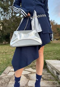 Personne portant une jupe asymétrique bleu marine, une veste rayée assortie et des chaussettes Adidas, tenant un sac à main Adidas argenté en plein air.