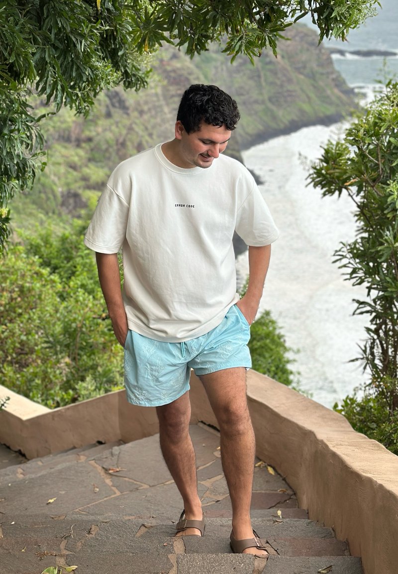 Giovane uomo che indossa una maglietta bianca con la scritta "ERROR CODE" e pantaloncini azzurri chiari che scende delle scale in pietra circondate da vegetazione, con oceano e scogli sullo sfondo.