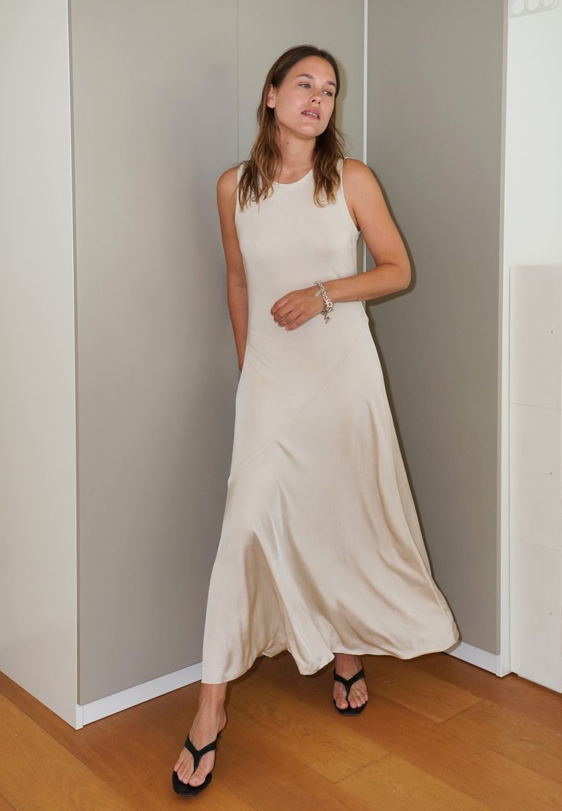 Beige ilmanhihaisen mekon, joka on valmistettu sileästä kankaasta ja jossa on epäsymmetrinen helma. Pukeutunut mustiin flip-floppeihin puulattialla.