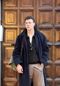 Jeune homme en pardessus foncé, pantalon beige, portant un sac vert, debout contre une porte en bois ornée, regardant sur le côté.