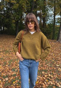 Oliivroheline kootud sviiter V-kaelusega, kombineeritud helesiniste teksadega. Pruneeritud pruuni käekotiga ja üle mõõtme päikeseprillidega. Sügisel lehestik taustal.